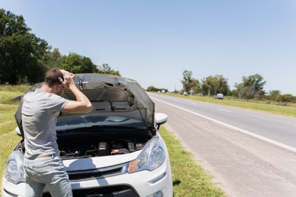 Frustrated man standing in front of a broken down car with its hood open on a rural road.