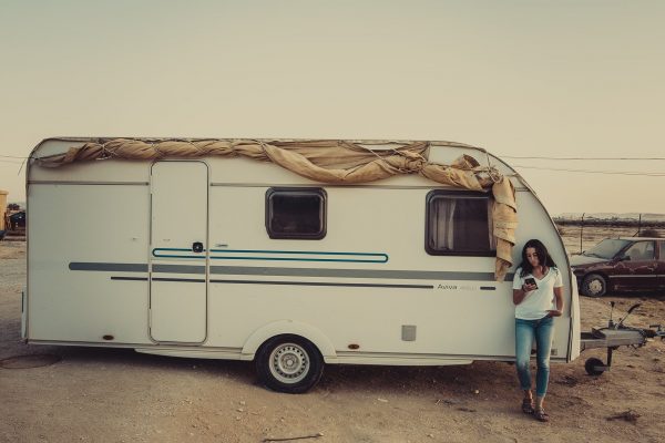 Woman standing next to a small, vintage white caravan in a dry, outdoor setting.